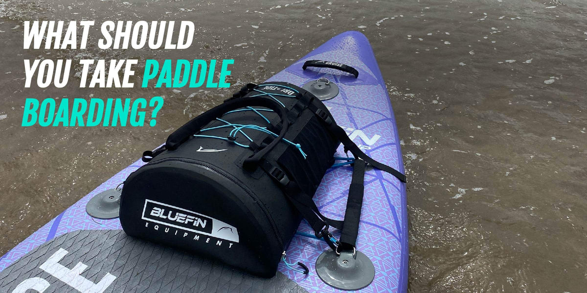 Paars paddleboard met zwarte uitrustingstas in ondiep water, met tekst over wat je meeneemt om te suppen