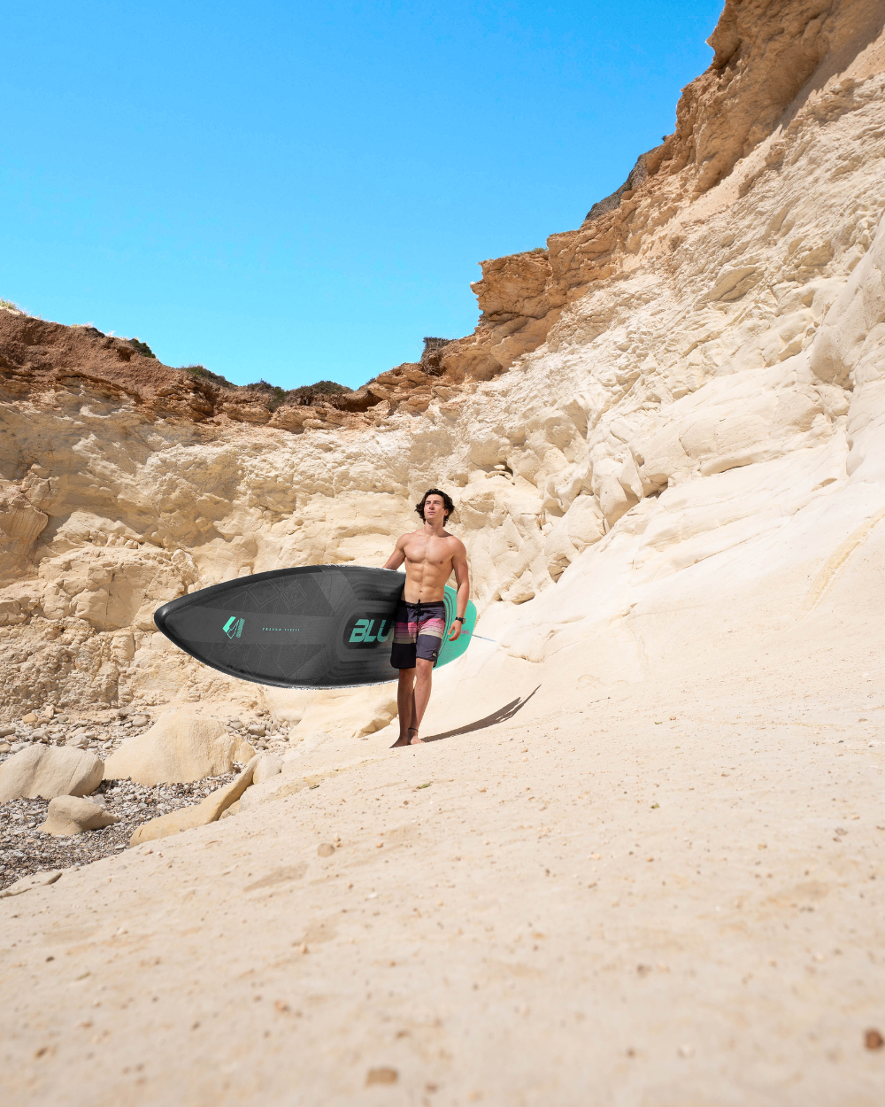 Man walking on sandy beach carrying a black paddleboard with green accents and rocky cliffs in background