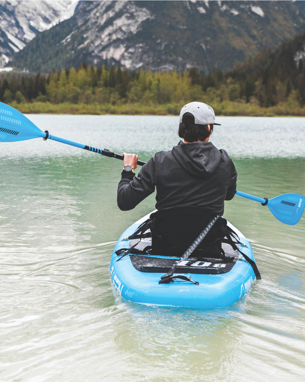 Person paddling a blue inflatable paddleboard on calm water with mountains in the background