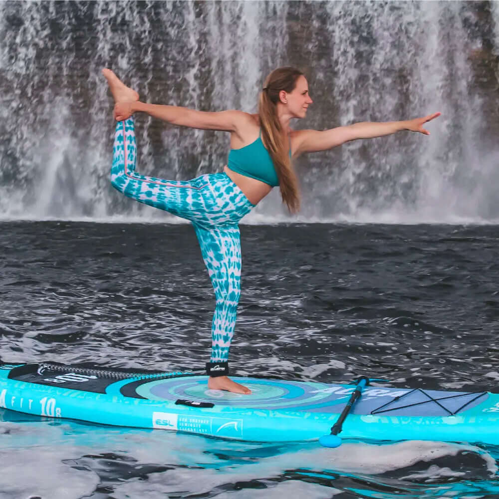 Woman in teal activewear performing yoga on a paddleboard in front of a waterfall