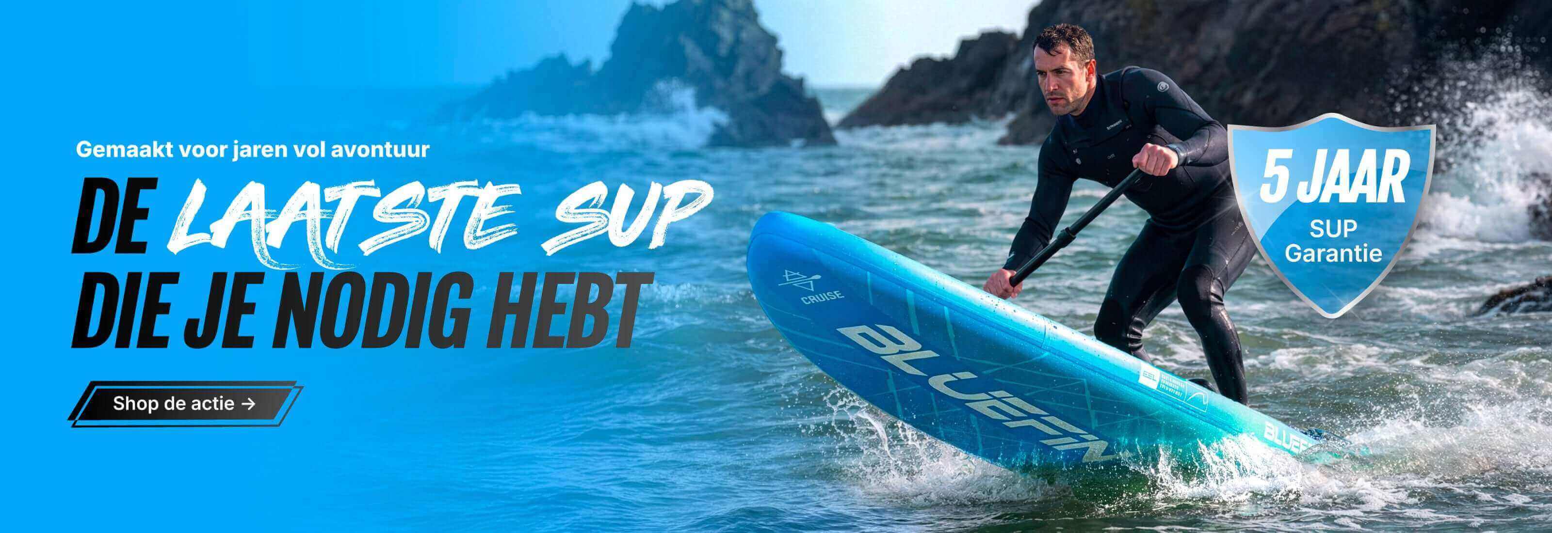 Man paddling a blue inflatable SUP on water, wearing a wetsuit, with rocky shore in background