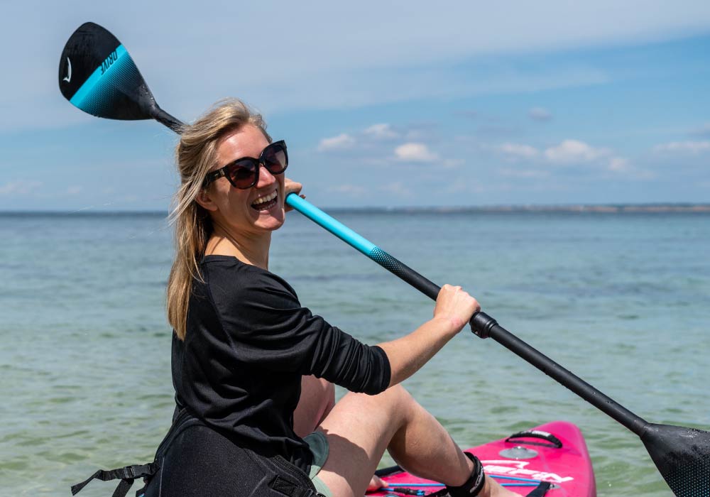 Woman smiling while sitting on a pink paddleboard, holding a blue paddle in shallow water.