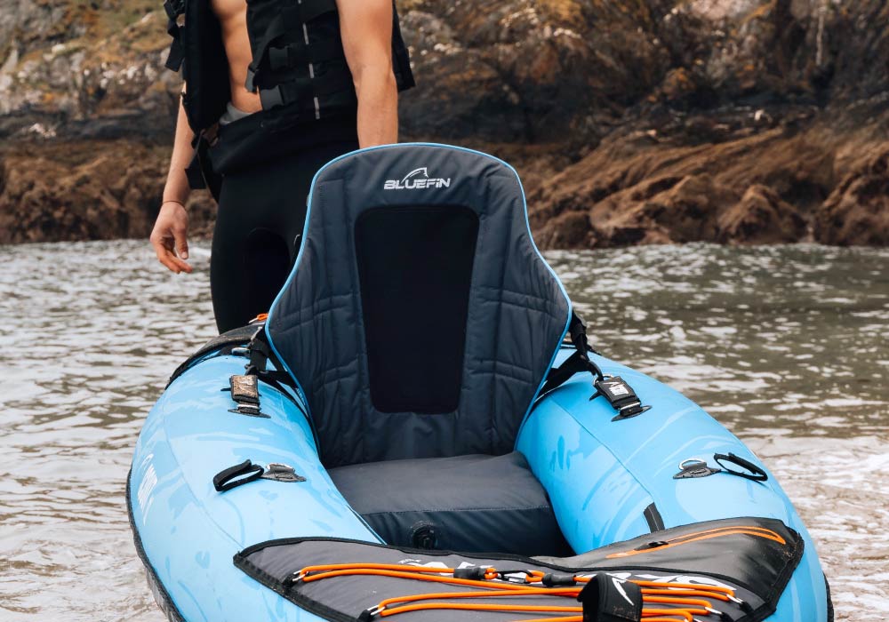 Blue inflatable kayak with a padded seat, near a rocky shore and a person standing beside it