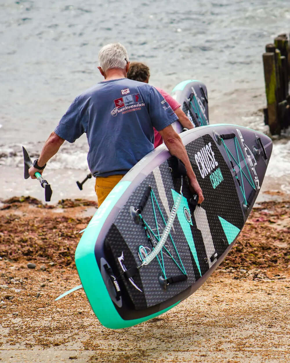 Two people carrying inflatable paddleboards towards the water, one with a paddle in hand.