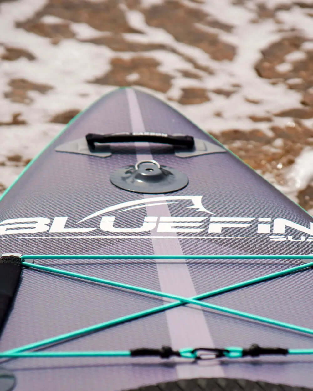 Inflatable paddleboard with Bluefin logo, bungee cords, and center fin on sandy shore