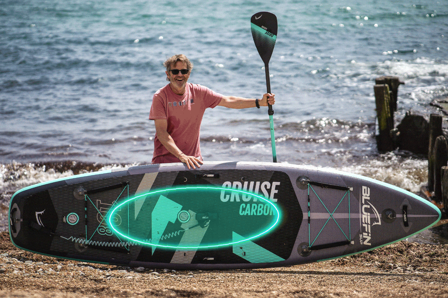 Man smiling beside a Bluefin inflatable paddleboard on a beach, holding a paddle.