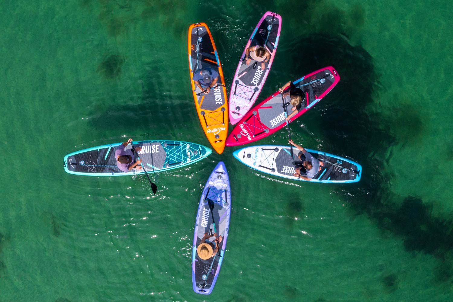 Six colorful paddleboards arranged in a circle on clear water, with paddlers using paddles.