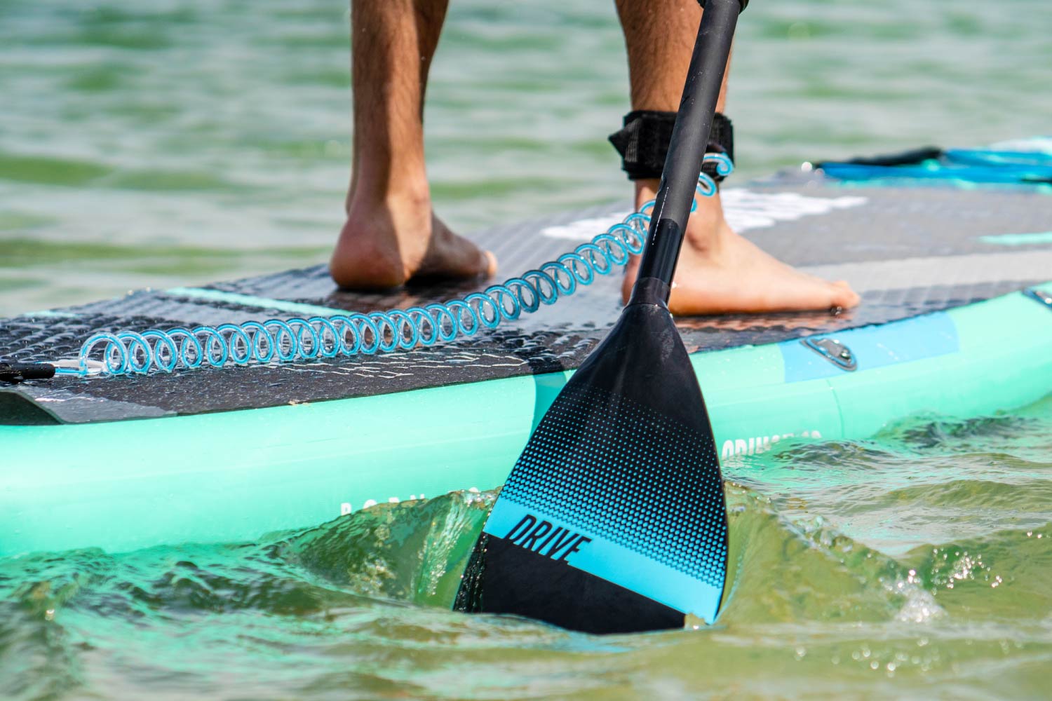 Person standing on a teal inflatable paddleboard, using a black paddle with blue accents in water.