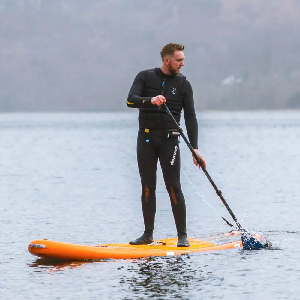 Man in wetsuit die peddelt op een oranje stand-up paddleboard op kalm water.