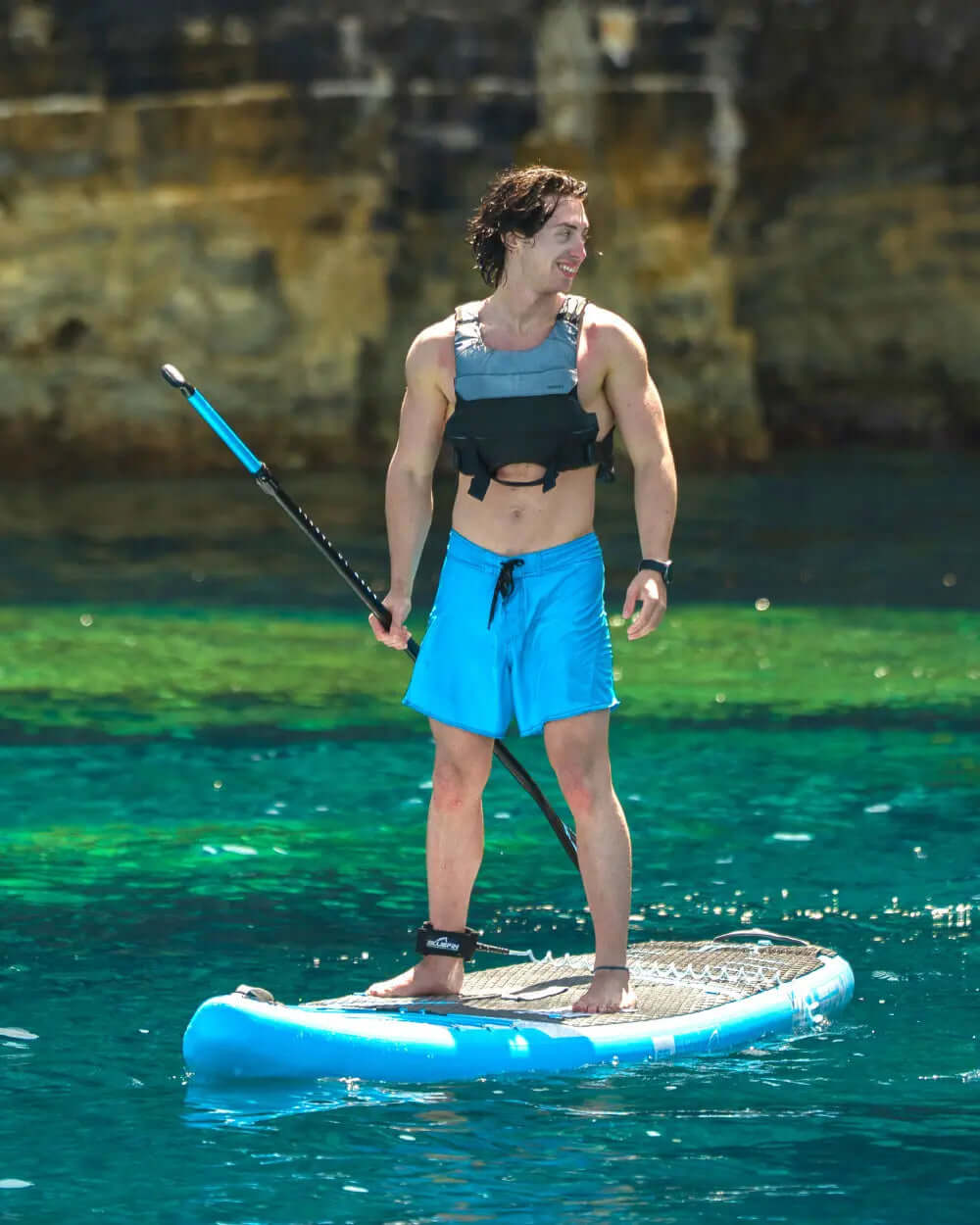 Man in a life vest and blue shorts paddleboarding on clear water with a paddle in hand