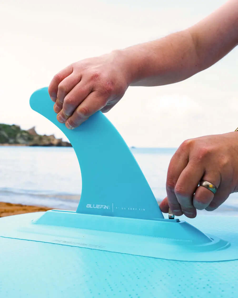 Person attaching a blue fin to a paddleboard on a beach with water in the background