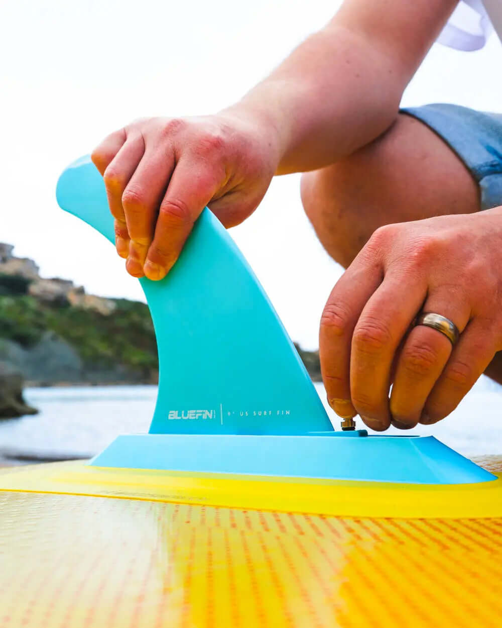 Person attaching a blue fin to a yellow paddleboard on a beach with rocky background