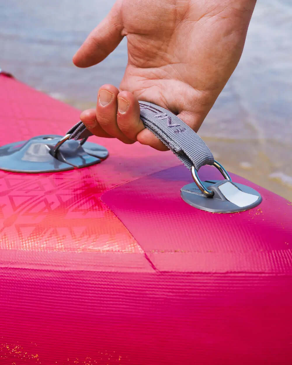 Hand gripping a gray strap attached to a pink inflatable paddleboard near the water