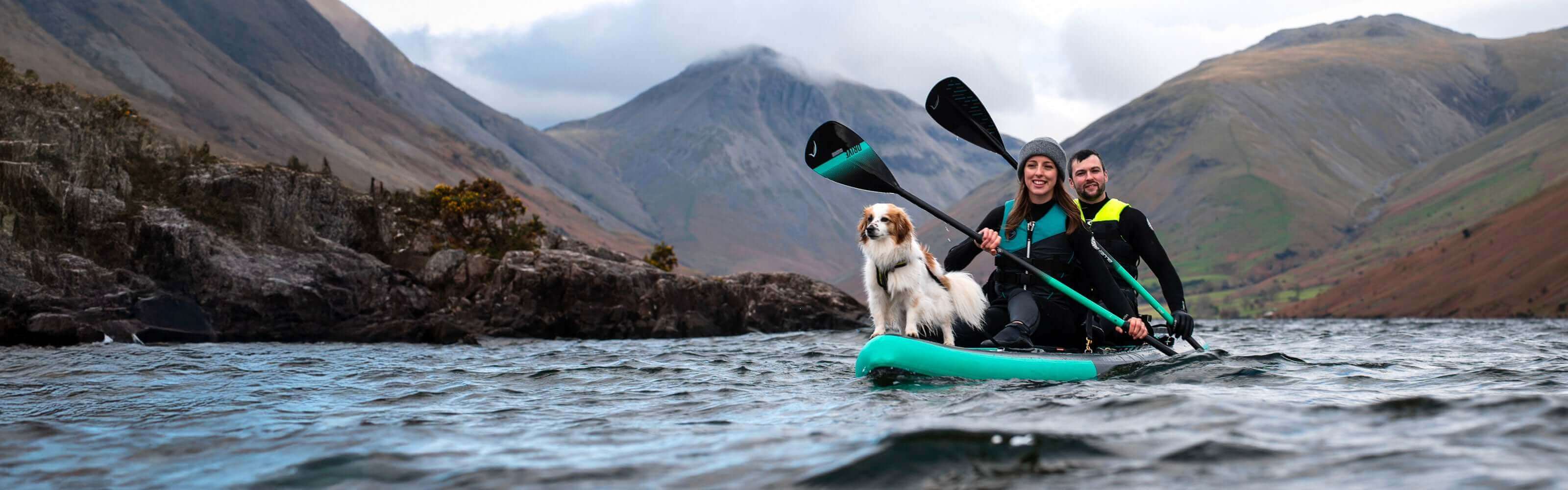 Two people and a dog on a teal inflatable paddleboard in a lake surrounded by mountains
