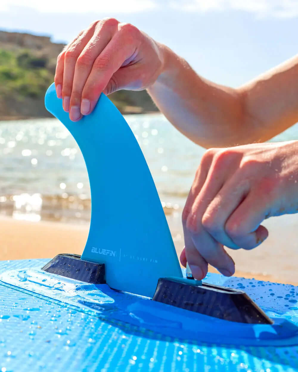 Person attaching a blue surf fin to a blue paddleboard on a sandy beach