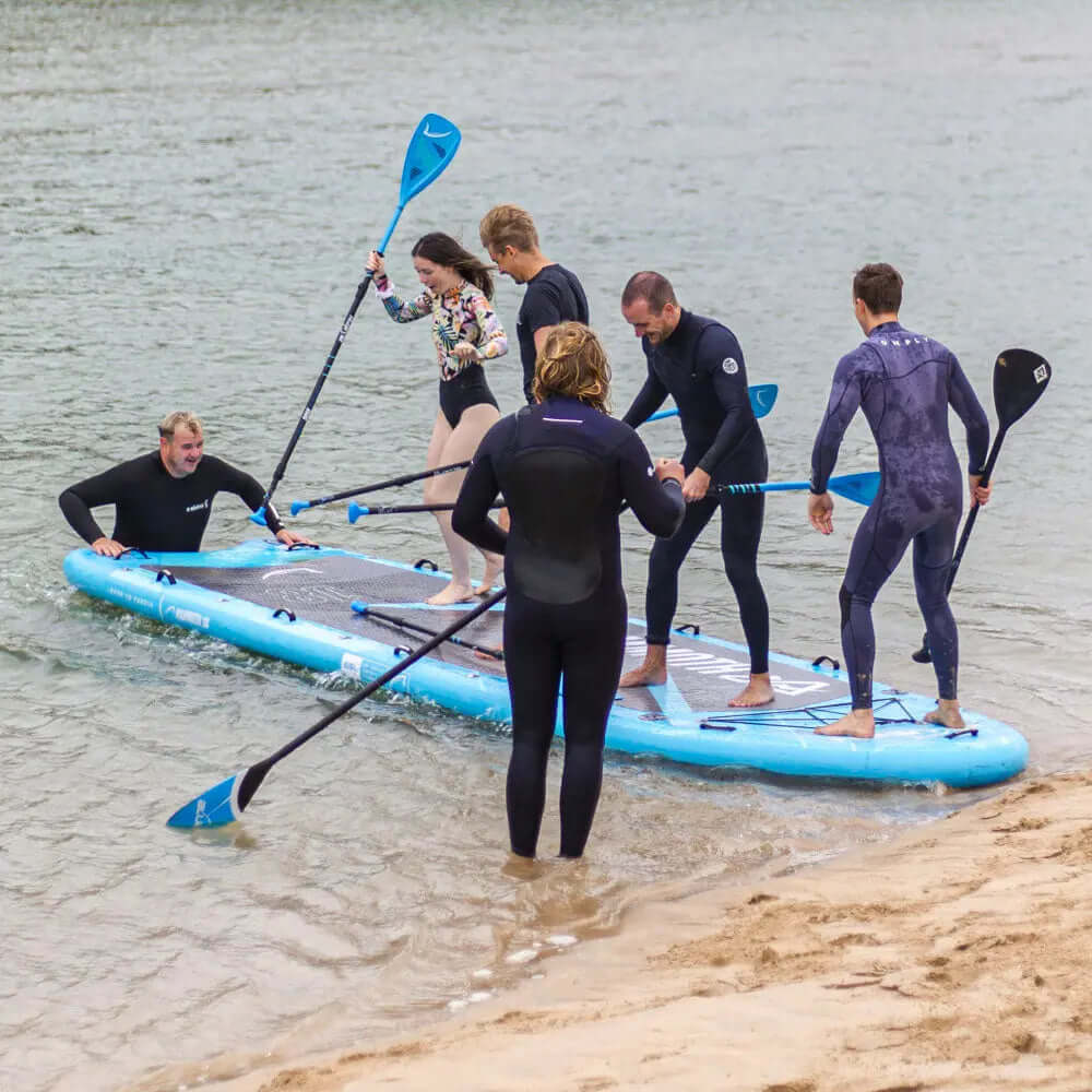 Groep van zes mensen op een groot blauw opblaasbaar paddleboard in ondiep water, sommigen peddelen, anderen balanceren.