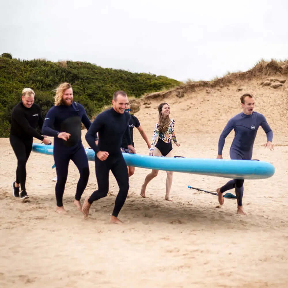 Groep van zes mensen in wetsuits die op zand rennen met een groot blauw paddleboard