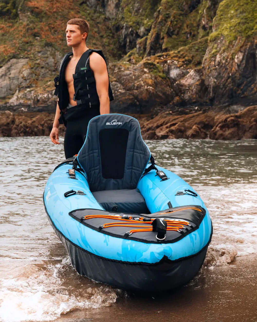 Inflatable kayak in blue with black accents, man in life jacket standing on sandy shore.
