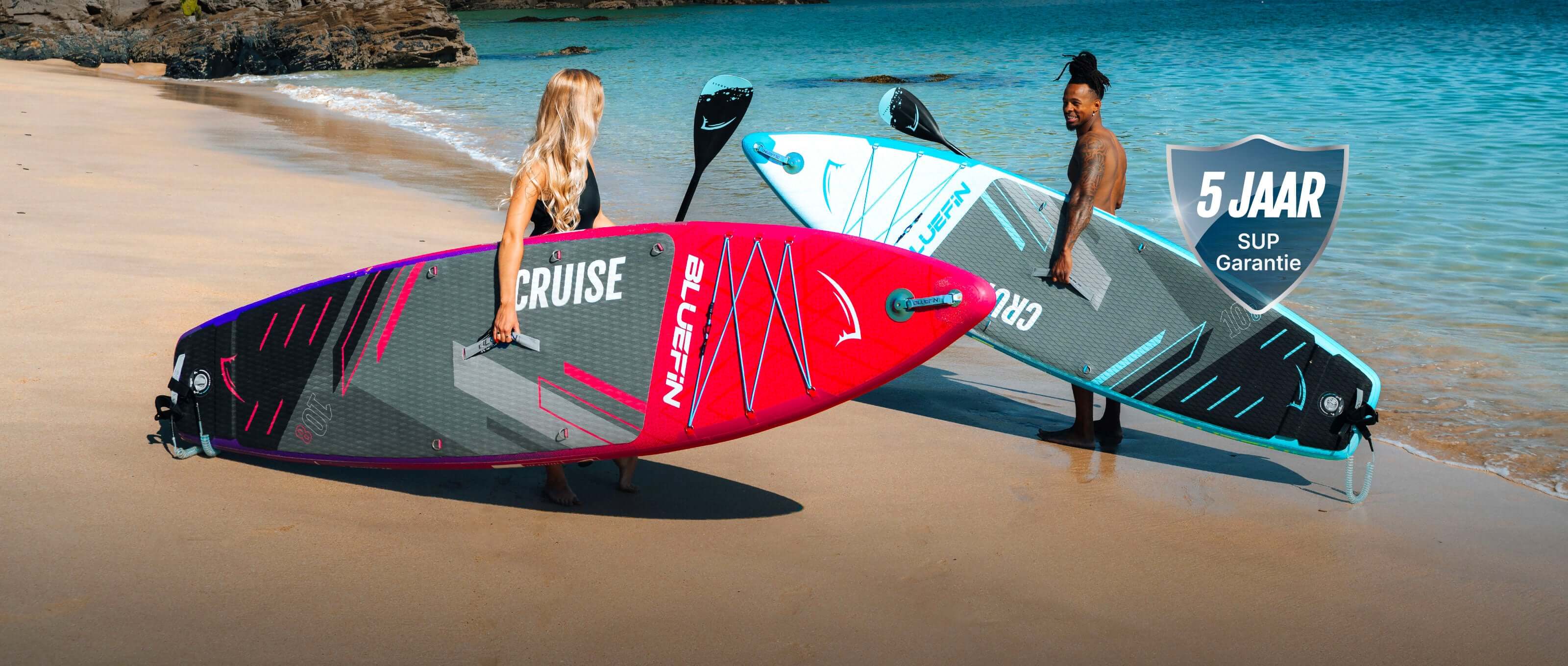 Two paddleboards on the beach, one pink and one blue, with two people holding them.