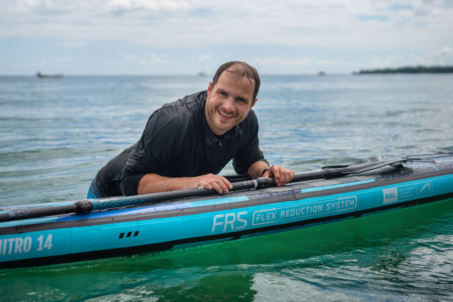 Man smiling while leaning on an inflatable paddleboard in calm water, with FRS branding visible