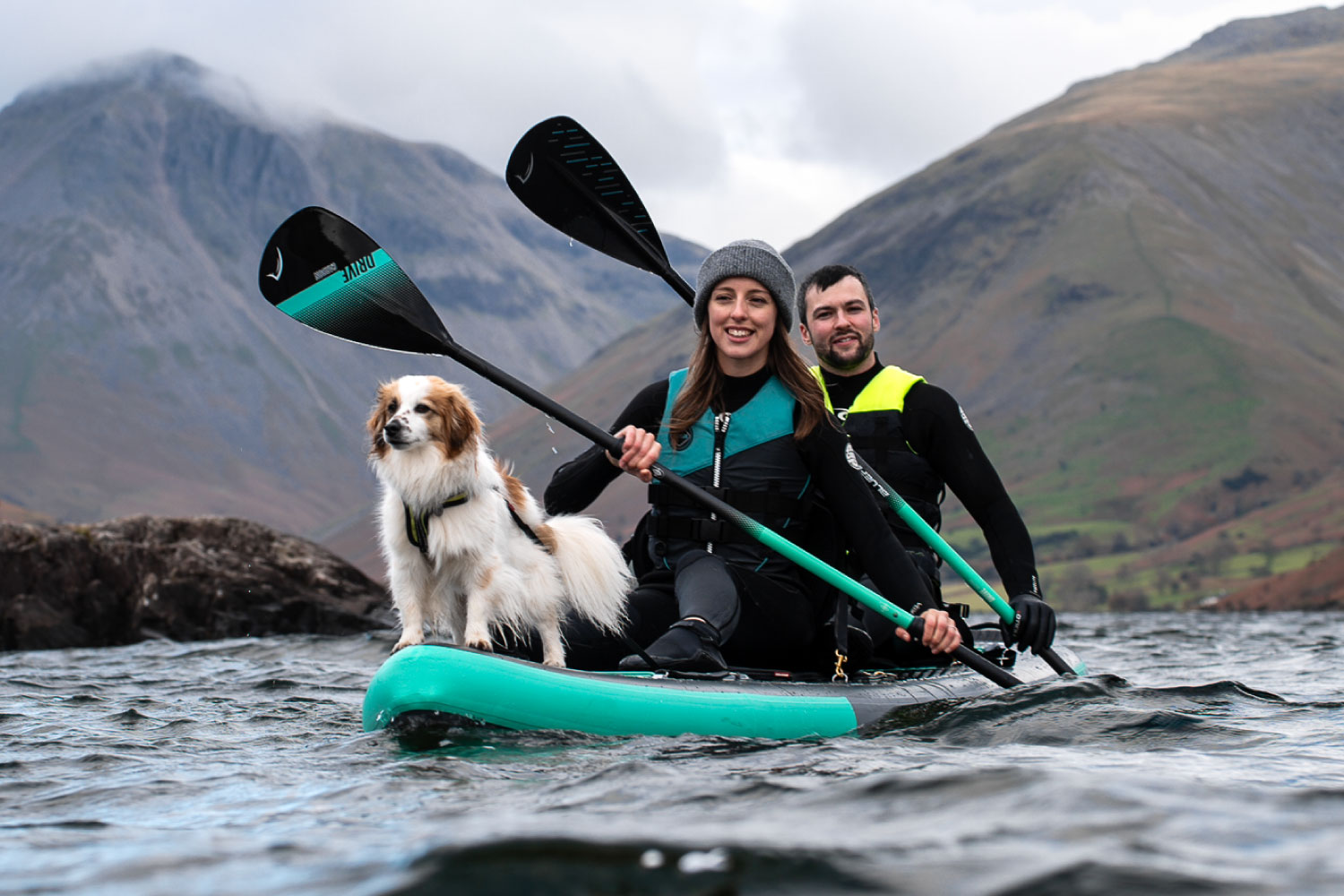 Two people and a dog on a teal inflatable paddleboard, paddling in a lake with mountains in the background.