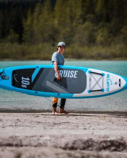 Person holding a Bluefin inflatable paddleboard by the water, wearing a cap and casual attire.
