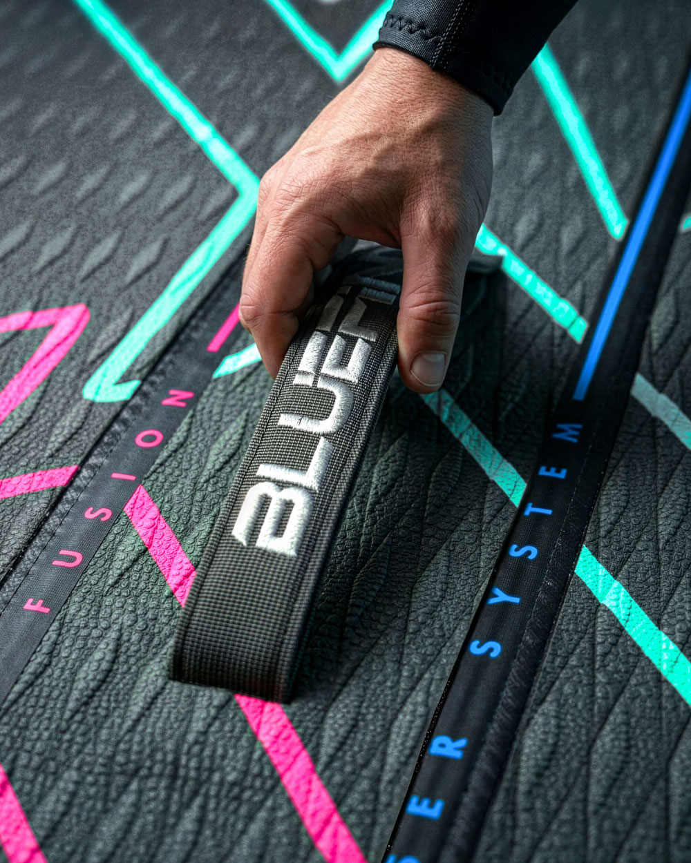 Hand gripping a textured strap labeled 'BLUEFIN' on a paddleboard with colorful graphics