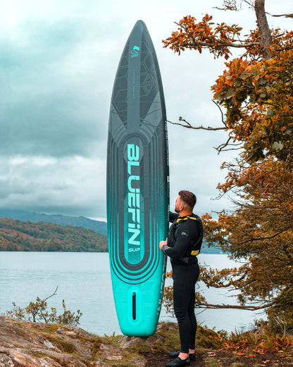 Inflatable Bluefin SUP paddleboard with textured deck, standing by a lake, person in wetsuit.