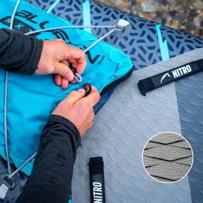 Handen die een blauwe opblaasbare paddleboard tas vastmaken met een grijze textuur op het dek en NITRO-riem.