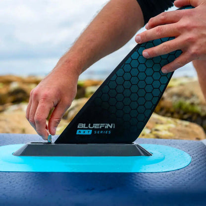 Persoon die een zwarte hexagonale vin bevestigt aan een Bluefin paddleboard op een rotsachtige kust.