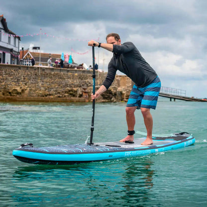 Man die peddelt op een blauw opblaasbaar paddleboard in kalm water, gekleed in een zwart shirt en gestreepte shorts.