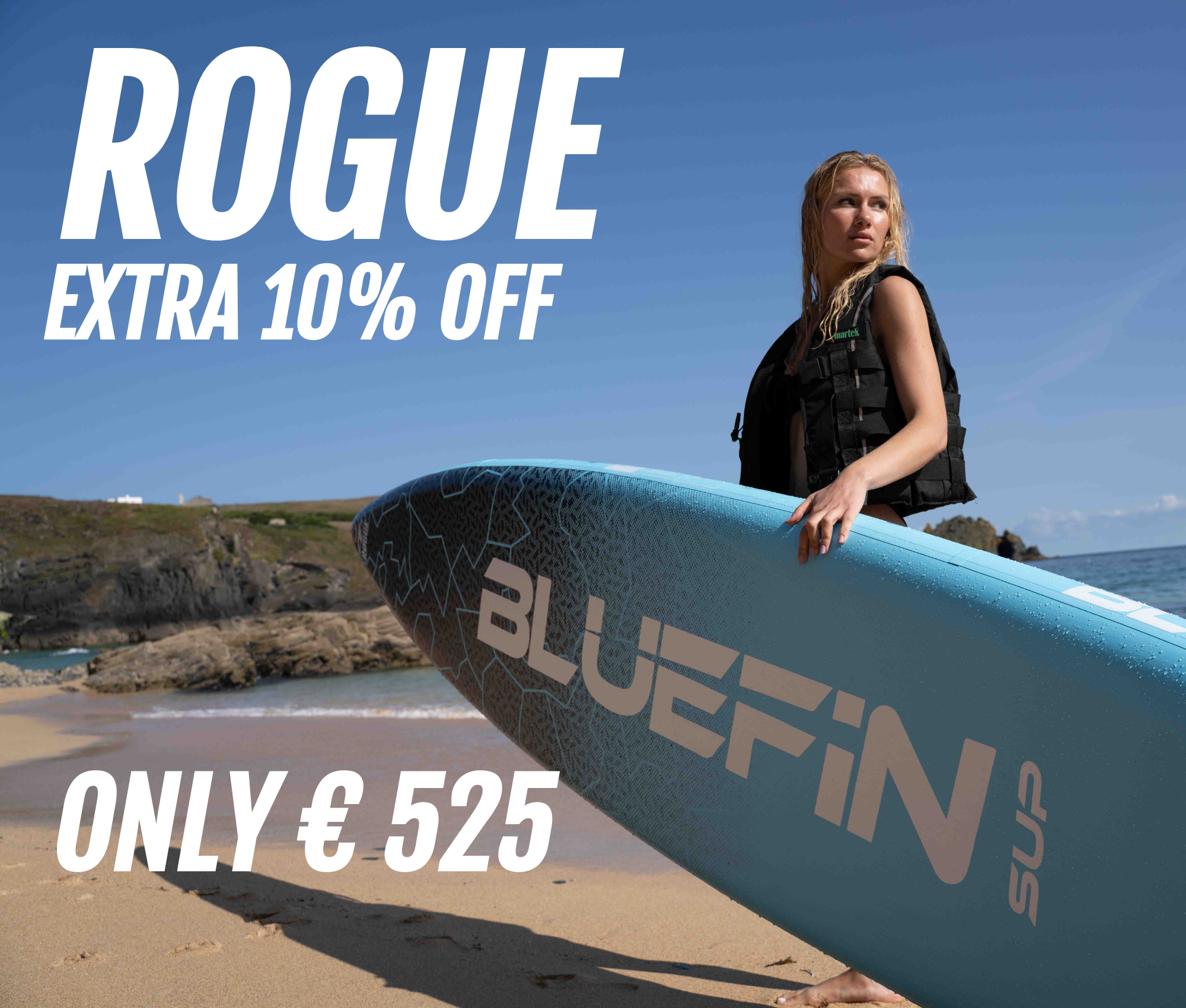 Woman holding a blue Bluefin SUP paddleboard on a sandy beach with rocky coastline