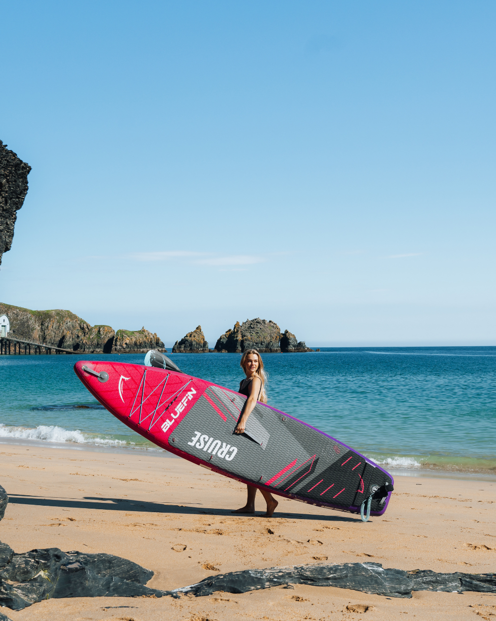 Woman holding a Bluefin inflatable paddleboard on a sandy beach with rocks and calm water in the background.