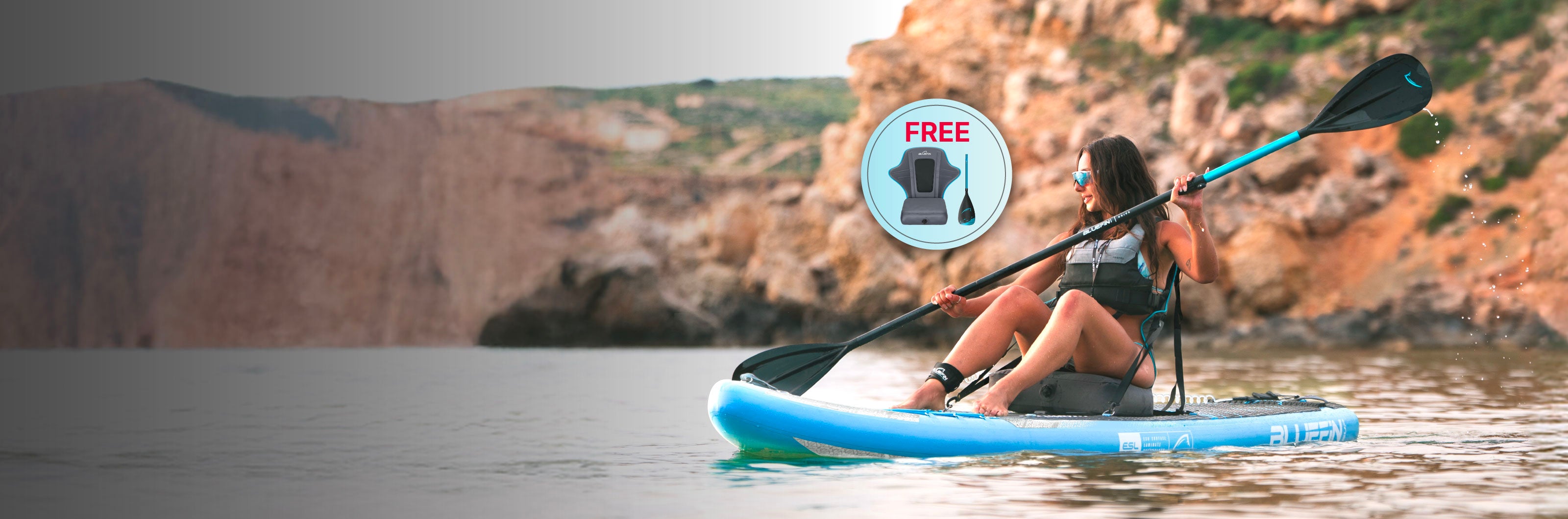Woman paddling on a blue inflatable paddleboard in calm water, rocky cliffs in background, free life vest offer visible